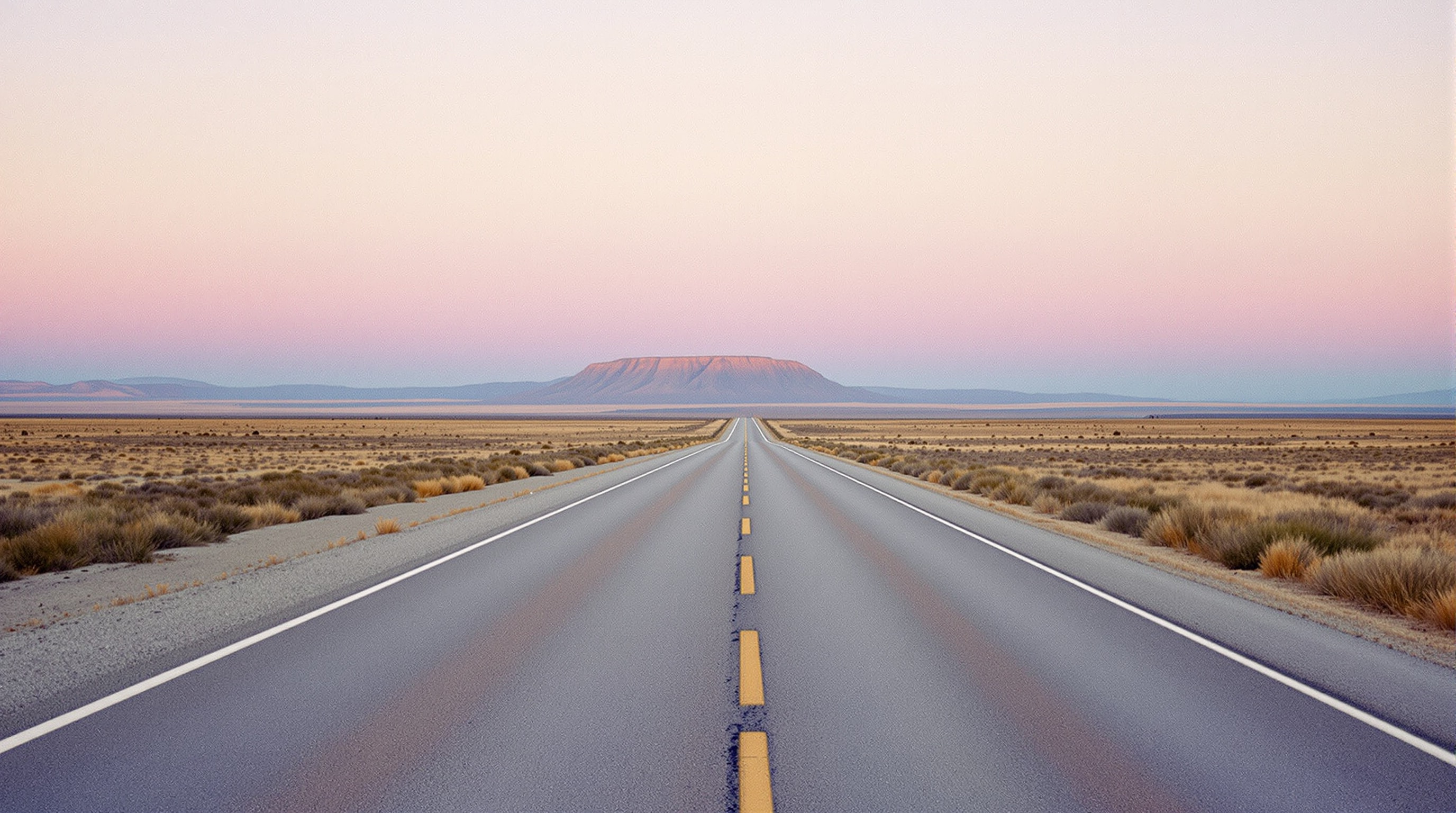 An empty desert highway running straight toward a distant mesa under a wide dawn sky